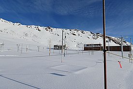 Eine weite Schneelandschaft mit einem Forschungsgebäude im Hintergrund. Im Vordergrund sind mehrere Stangen und Messinstrumente, die zur meteorologischen oder wissenschaftlichen Datensammlung verwendet werden. Die Umgebung wirkt unberührt und ruhig, umgeben von Bergen unter einem klaren Himmel.