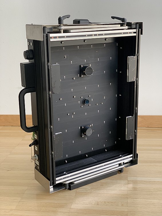 SnowImager Gerät mit schwarzer Oberfläche, drei zentralen Knöpfen und Metallrahmen, auf Holzboden liegend