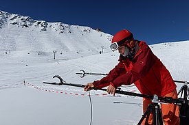 Anja Mödl in rotem Helm und rotem Skianzug installiert eine Messstation im Schnee vor schneebedeckten Bergen unter klarem blauem Himmel.