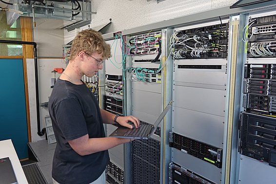 Ein junger Mann steht vor einem Serverraum und arbeitet mit einem Laptop. Hinter ihm sind mehrere Server mit sichtbaren Kabeln und Komponenten angeordnet. Die Umgebung wirkt technisch und modern.