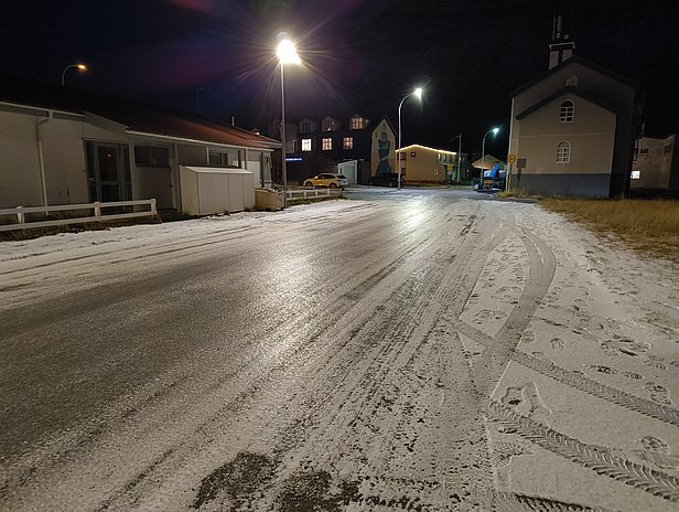 Die Aufnahme zeigt eine schneebedeckte Straße bei Nacht, beleuchtet von Straßenlampen. Auf der linken Seite befinden sich einfache Häuser, während im Hintergrund einige Gebäude und ein Kirchengebäude sichtbar sind. Die Atmosphäre wirkt ruhig und den winterlichen Bedingungen entsprechend.