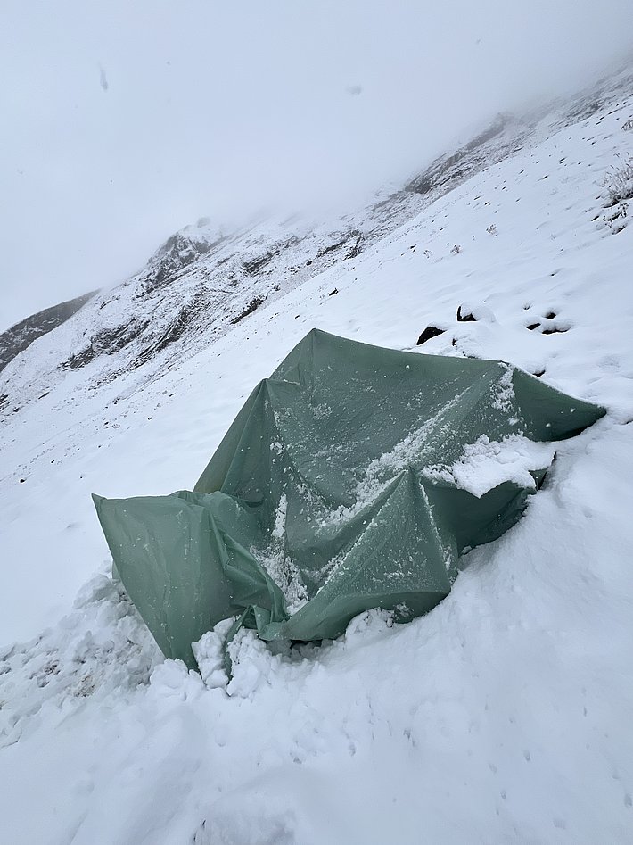Ein grüne Abdeckplane liegt in einer schneebedeckten Landschaft. Der Himmel ist bewölkt und die Umgebung wirkt kalt und rau. Schnee ist auf der Plane und dem Boden sichtbar.