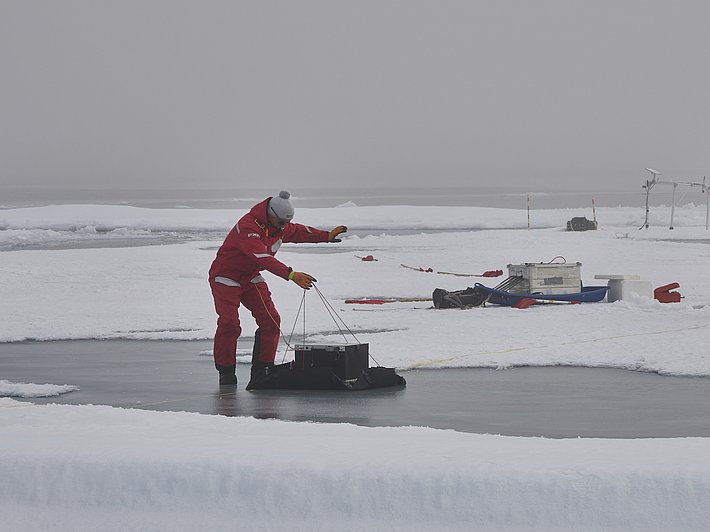 Ein Mann in roter Winterkleidung und grauer Mütze steht auf einer gefrorenen Fläche und zieht ein schwarzes Gerät mit einem Seil. Im Hintergrund sind weitere Ausrüstungsgegenstände auf dem Eis.