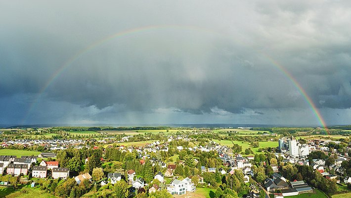A rainbow arches over a green, sun-drenched landscape with a town in the foreground. The sky is gloomy and covered in dark clouds, but in places the sun shines through, creating a contrast to the overcast atmosphere.