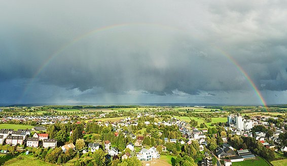 Un arcobaleno si estende su un paesaggio verde e soleggiato, con una città in primo piano. Il cielo è cupo e coperto da nuvole scure, ma in alcuni punti il sole fa capolino, creando un contrasto con l'atmosfera nuvolosa.