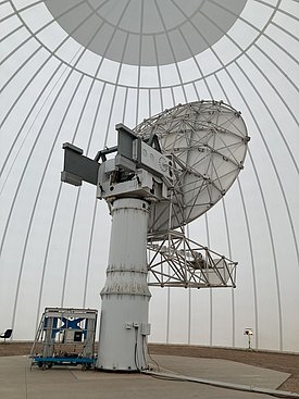 Ein großer Satellitenschüssel-Mast steht in einer lichtdurchfluteten Kuppel. Die Schüssel ist auf einem stabilen, zylindrischen Sockel montiert und ist an einem beweglichen Steuerungsmechanismus befestigt.