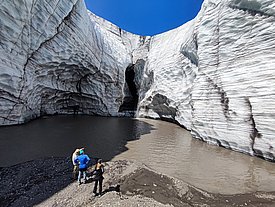 Three people stand on the shore of a meltwater lake in front of tall, layered glacier walls with a dark cave in the center under a blue sky.