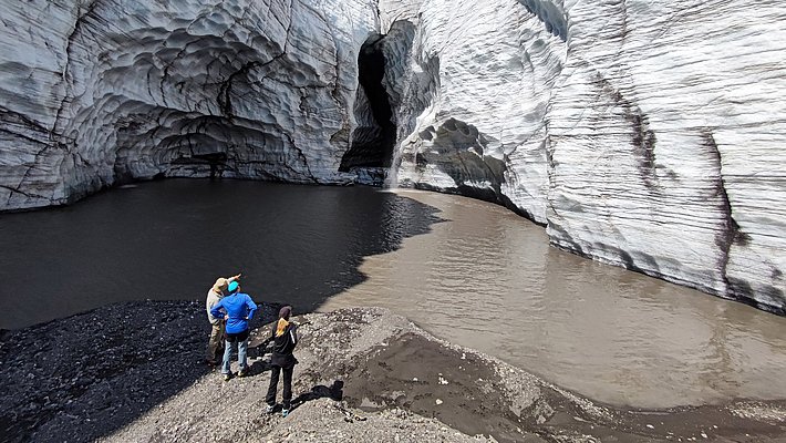 Drei Personen stehen am Ufer eines Schmelzwassersees vor hohen, geschichteten Gletscherwänden mit einer dunklen Höhle in der Mitte unter blauem Himmel.