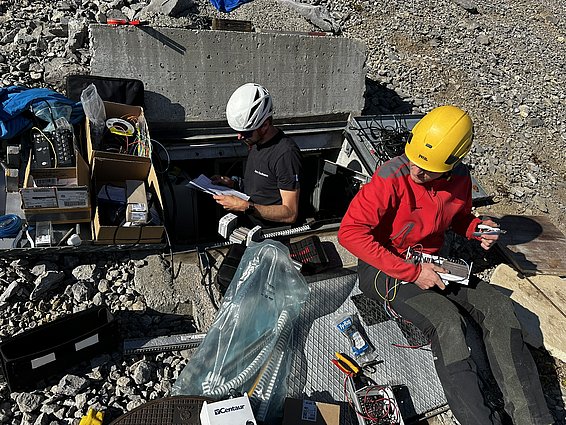 Zwei Personen arbeiten im Freien an technischen Geräten. Eine Person sitzt mit Werkzeug und Kabeln auf dem Boden, während die andere in einem offenen Schacht sitzt und eine Anleitung liest. Um sie herum sind verschiedene Kabel und Kisten verteilt.