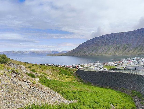 Eine panoramische Ansicht zeigt eine ruhige Bucht, umgeben von grünen Hügeln und einer bewaldeten Flussmündung. Im Hintergrund erhebt sich ein steiler Berg, während am Ufer eine kleine Stadt mit bunten Häusern sichtbar ist, die einen malerischen Eindruck hinterlässt.
