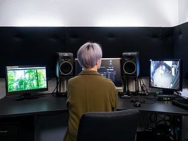 Person mit kurzen, hellvioletten Haaren sitzt vor drei Monitoren in einem schallisolierten Raum, zwei Lautsprecher stehen links und rechts der Monitore.