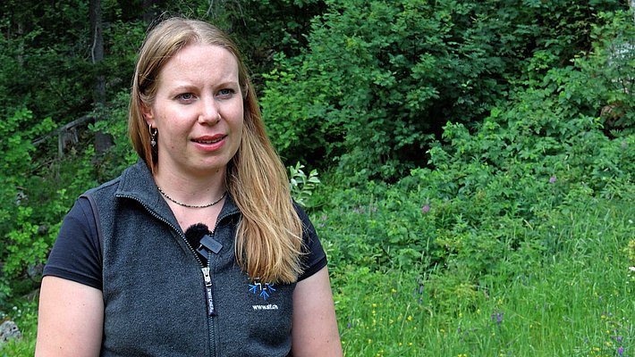 Woman with long blonde hair wearing a black t-shirt standing in front of dense forest