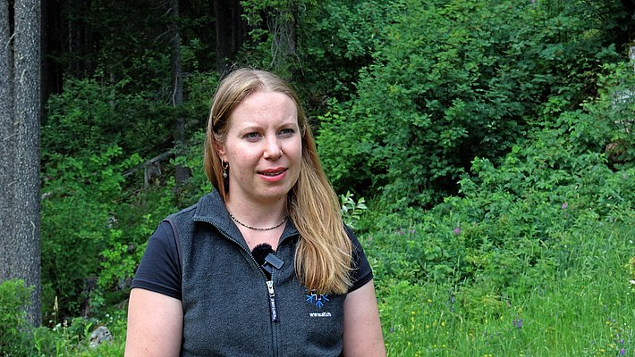 Frau mit langen blonden Haaren und schwarzem T-Shirt vor dichtem Wald