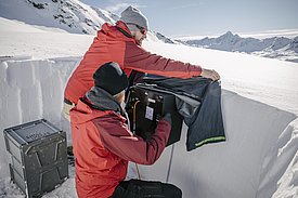 Zwei Männer in roten Jacken führen einen Feldversuche mit einem Prototyp des SnowImagers durch in einer Schneewand in einer verschneiten Berglandschaft.