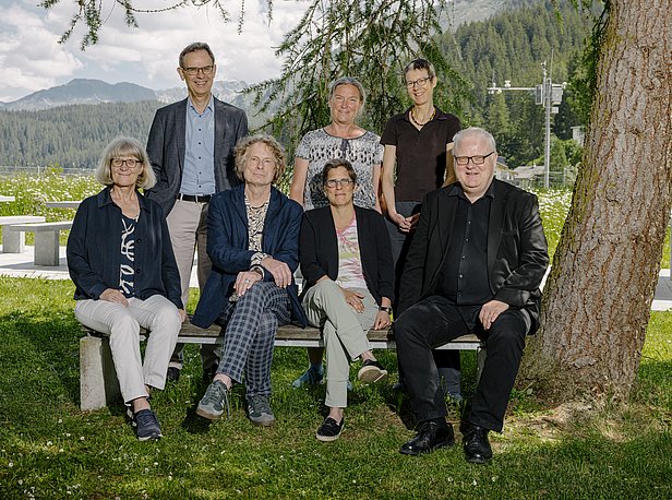 Eine Gruppe von sieben Personen sitzt auf einer Bank im Freien, umgeben von Bäumen und einer Landschaft mit Bergen im Hintergrund. Es sind sowohl Frauen als auch Männer unterschiedlichen Alters zu sehen, die freundlich in die Kamera schauen.