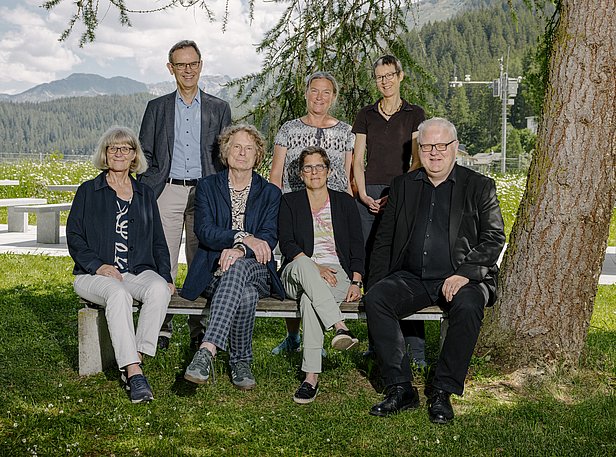 Eine Gruppe von sieben Personen sitzt auf einer Bank im Freien, umgeben von Bäumen und einer Landschaft mit Bergen im Hintergrund. Es sind sowohl Frauen als auch Männer unterschiedlichen Alters zu sehen, die freundlich in die Kamera schauen.