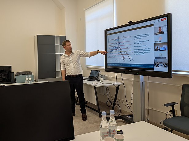 Mann in weißem Hemd zeigt auf großen Bildschirm mit Diagramm und Text in hellem Büro mit Schreibtisch und Stühlen.
