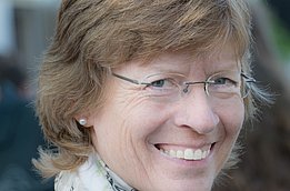 Femme aux cheveux blonds courts, portant des boucles d'oreilles en perle et un foulard clair à motif floral.