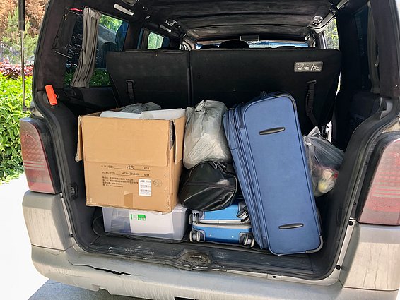 The image shows the trunk of a vehicle filled with various items, including a large cardboard box, several pieces of luggage, and bags. The contents are packed closely together, indicating preparation for a trip.
