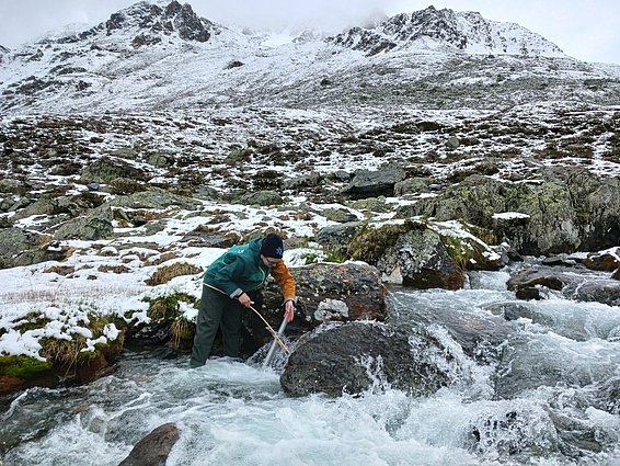 Winter mountain landscape with Maria in the foreground, accessing one of her river water temperature sensors. 
