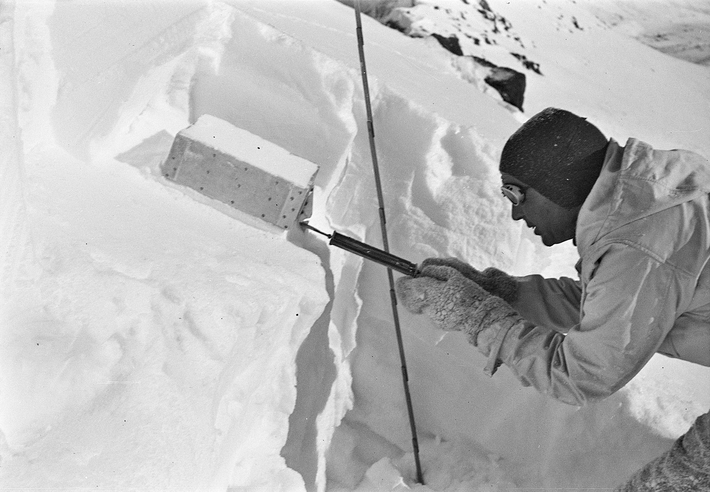 A man dressed warmly with a hat, gloves and glacier goggles takes a snow sample with a shear apparatus.