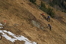 Drei Personen installieren Sensoren an einem steilen, trockenen Hang mit vereinzeltem Schnee und Nadelbäumen im Hintergrund.
