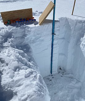 Ausgegrabener Schneekern mit sichtbaren Schneeschichten und einem blauen Messstab zur Temperaturmessung, daneben stecken Probenfläschchen im Schnee