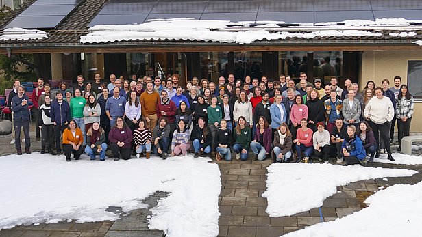 A large group of diverse individuals, comprising both men and women of various ages, is gathered outdoors in front of a building with a snow-covered ground. They stand closely together, smiling, wearing casual clothing, with some wearing name tags, indicating a friendly gathering or event.