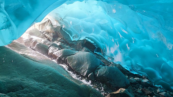 A view from inside an ice cave with smooth, bluish ice walls and rocks on the floor