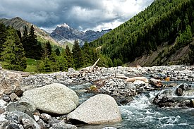 A rushing river flows over smooth stones, surrounded by a lush green forest and steep mountains. Dark clouds gather above, hinting at possible rain. The landscape conveys a tranquil yet dramatic natural environment.