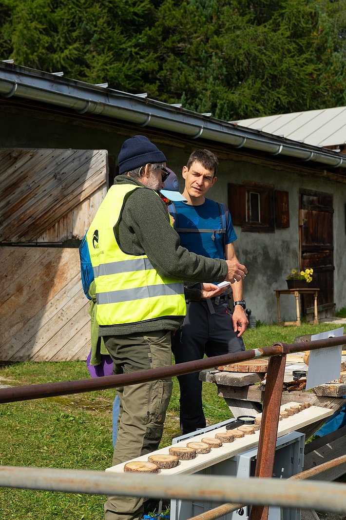 Eine Person mit grauem Bart trägt eine gelbe Warnweste und einen blauen Hut. Sie steht neben einer Person in gestreiftem Shirt. Im Vordergrund sind Holzscheiben auf einem Tisch angeordnet.