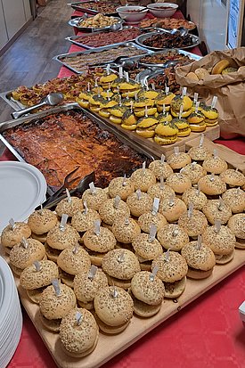 Ein gedeckter Tisch mit verschiedenen Speisen, darunter verschiedene Kuchen, Gebäck und Salate. Eine große Auswahl an Snacks und Süßigkeiten ist auf mehreren Platten angerichtet, die einladend aussehen. Im Hintergrund ist eine Tür zu sehen, die nach draußen führt.