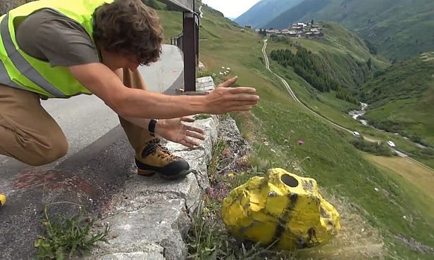 Ein Forscher in Leuchtweste lässt einen gelb angemalten, mit Sensoren bestückten Stein über eine Strassenkante einen Berghang hinunterrollen. 