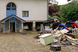 Auf einem schlammbedeckten Vorplatz eines Hauses stapeln sich mehrere Autos, Bretter und vom Hochwasser versehrter Hausrat.