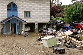 Auf einem schlammbedeckten Vorplatz eines Hauses stapeln sich mehrere Autos, Bretter und vom Hochwasser versehrter Hausrat.