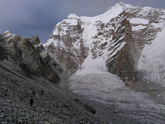 Three hikers ascend a rocky slope towards a towering snow-covered mountain. The landscape features steep, rugged terrain, with glaciers reflecting the muted light of an overcast sky. The scene conveys the challenges and beauty of mountainous exploration.