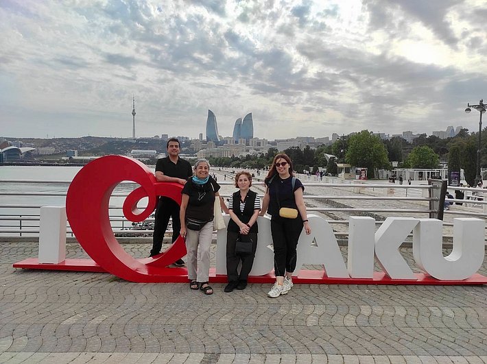 Four people pose in front of large red lettering reading ‘I ❤️ Baku’ on the shore. The cityscape with modern buildings is visible in the background. The sky is partly cloudy.