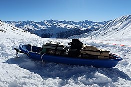 Blauer Schlitten mit Messgeräten und Ausrüstung auf schneebedecktem Bergplateau, umgeben von schneebedeckten Bergen unter klarem Himmel