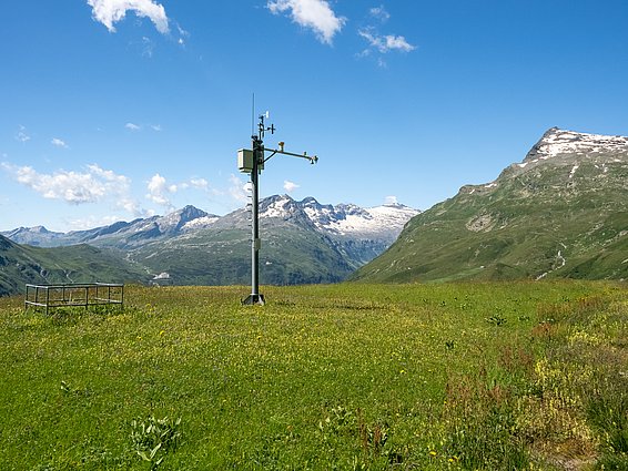 Messstation auf einer grünen Bergwiese mit schneebedeckten Bergen im Hintergrund unter blauem Himmel