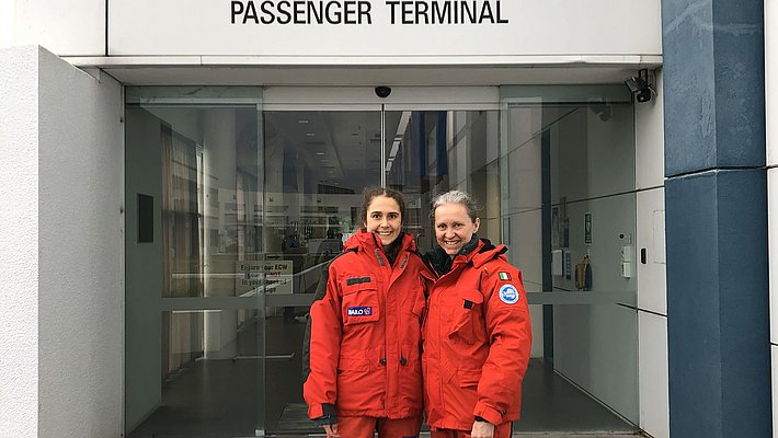Due persone in tute da spedizione arancioni sono in piedi davanti all'ingresso del terminal passeggeri dello United States Antarctic Program a Christchurch.