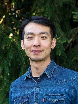 Porträt eines Mannes mit kurzem, dunklem Haar, der in einem blauen Jeanshemd vor einem Hintergrund aus grünen Pflanzen steht. Er lächelt freundlich in die Kamera.