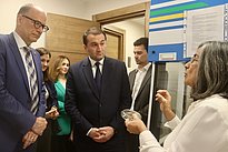 A group of professionals, including a woman in a white lab coat explaining something, gathers in a room. Two men in formal attire listen attentively.