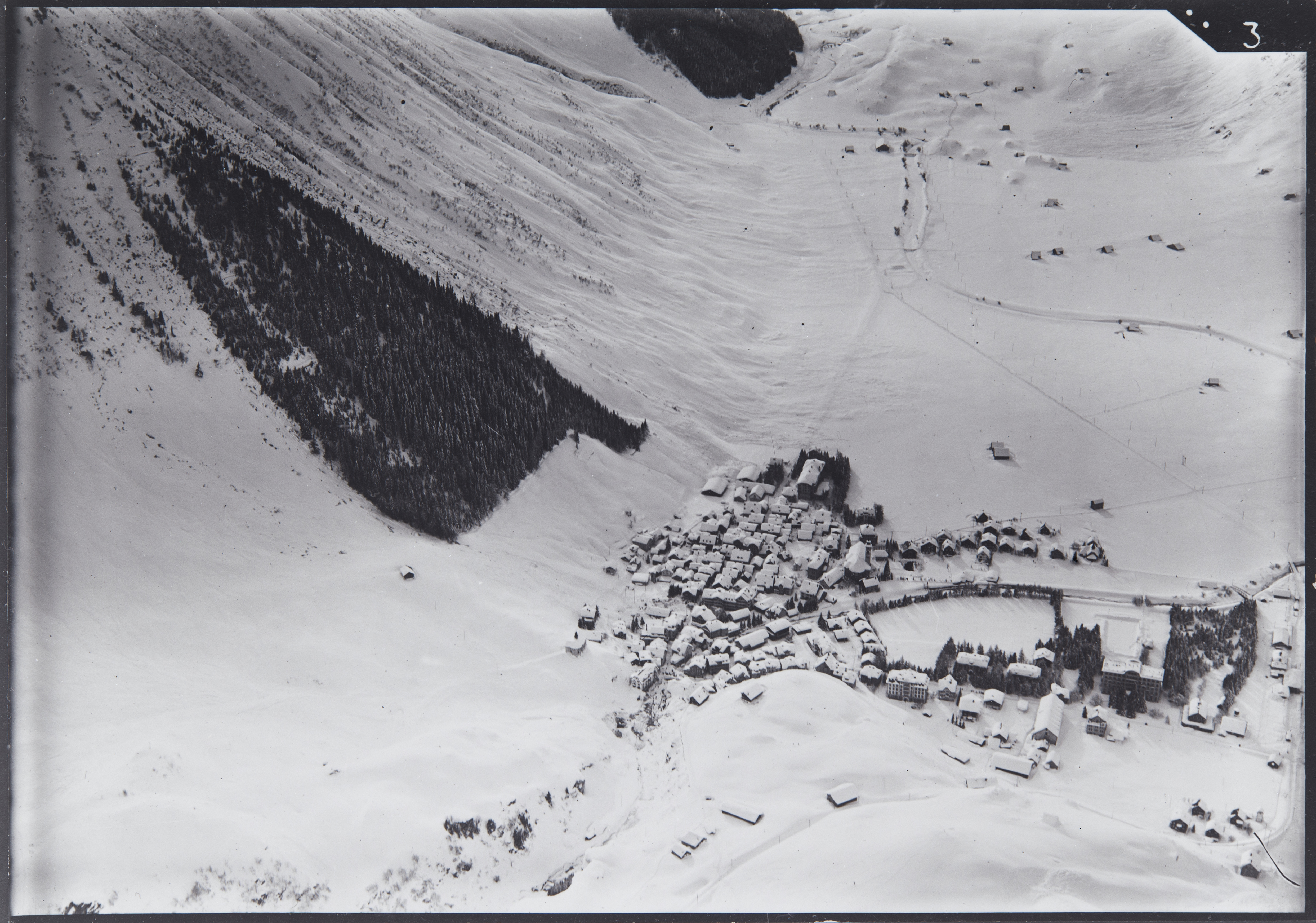 Black and white aerial photograph of a snow-covered mountain village with forest on the hillside.