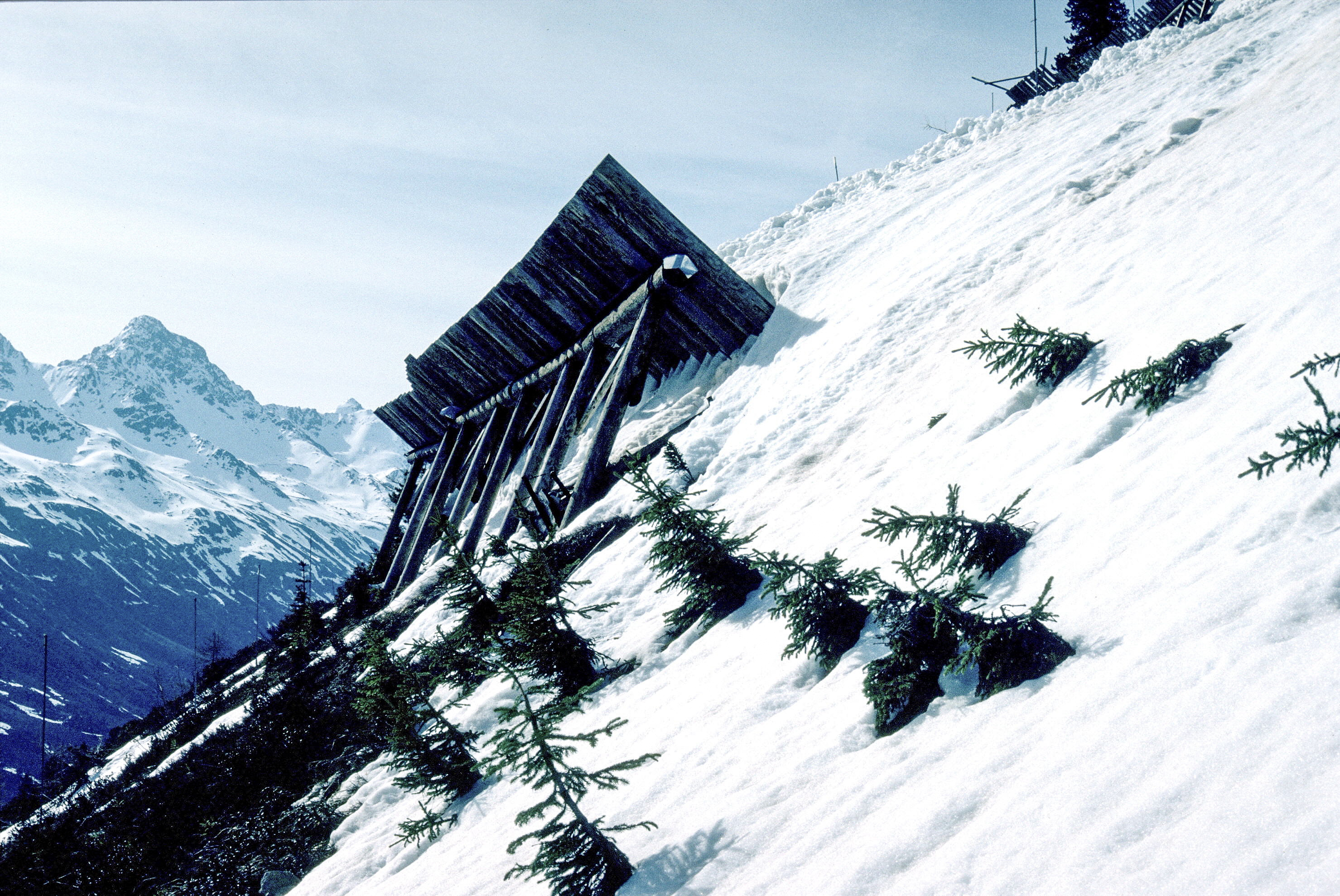 Schneefangzaun aus Holz auf schneebedecktem Berghang mit Tannen und Berggipfeln im Hintergrund.